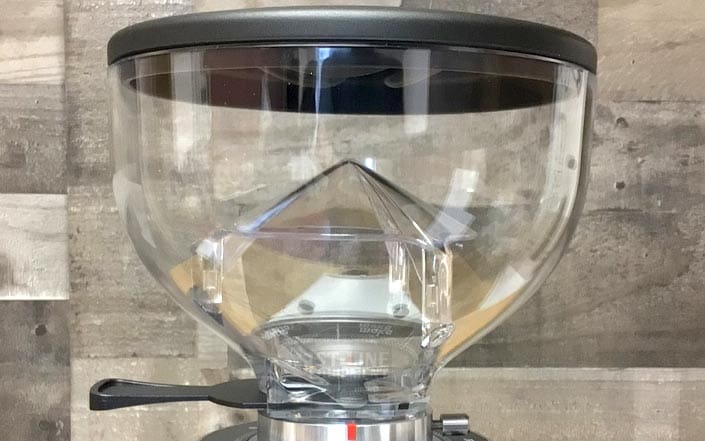 A close-up of the Fiorenzato AllGround Coffee Grinder - Time-Based Grinding shows its transparent hopper with a black lid, mounted on the grinder against a wood-patterned background. Ideal for precise, timed grinding. © 2025 1st-line.com