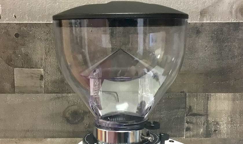 A close-up of the transparent hopper with a black lid on the Quamar M80TP Espresso Coffee Grinder, set against a wooden textured wall. © 2025 1st-line.com