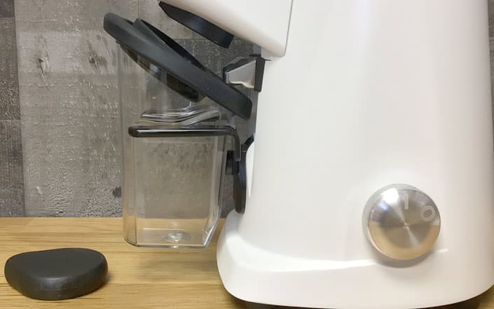 A close-up side view of the Fiorenzato AllGround Coffee Grinder - Time-Based Grinding, featuring a clear container, large silver knob, and black lid on a wooden surface with a gray textured wall in the background. © 2025 1st-line.com