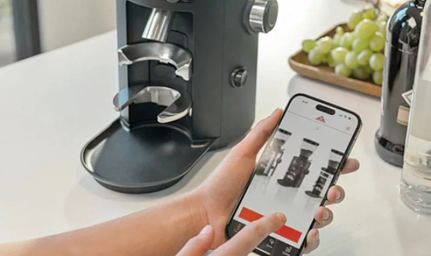A person uses a smartphone app in front of a black Mahlkonig X54 Allround Home Grinder on a kitchen counter, with green grapes and a wine bottle in the background. © 2025 1st-line.com