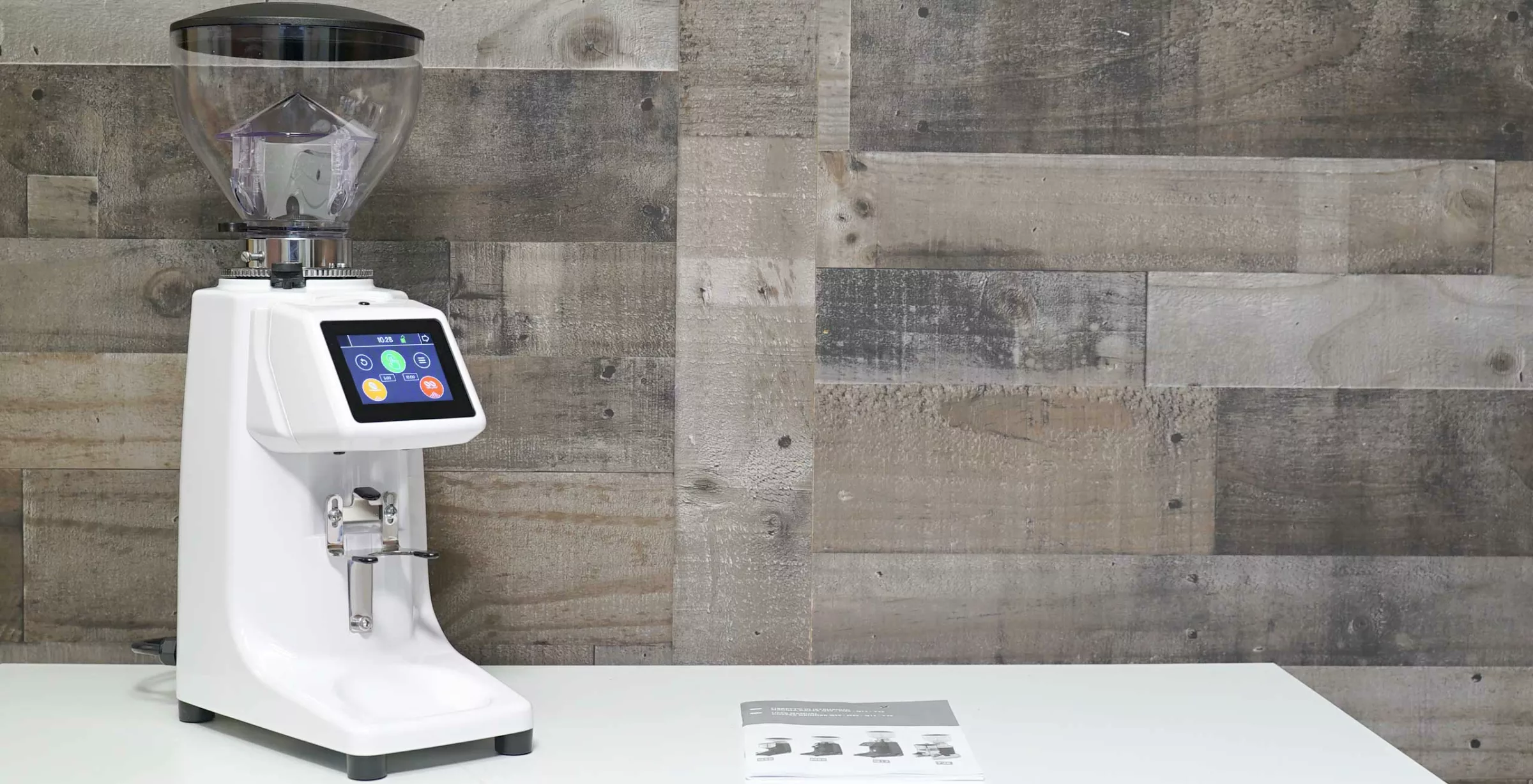 A Quamar M80TP Espresso Coffee Grinder in white, featuring a touchscreen display, rests on a white surface before a wooden plank wall. An instruction manual is placed to the right of the grinder. © 2025 1st-line.com