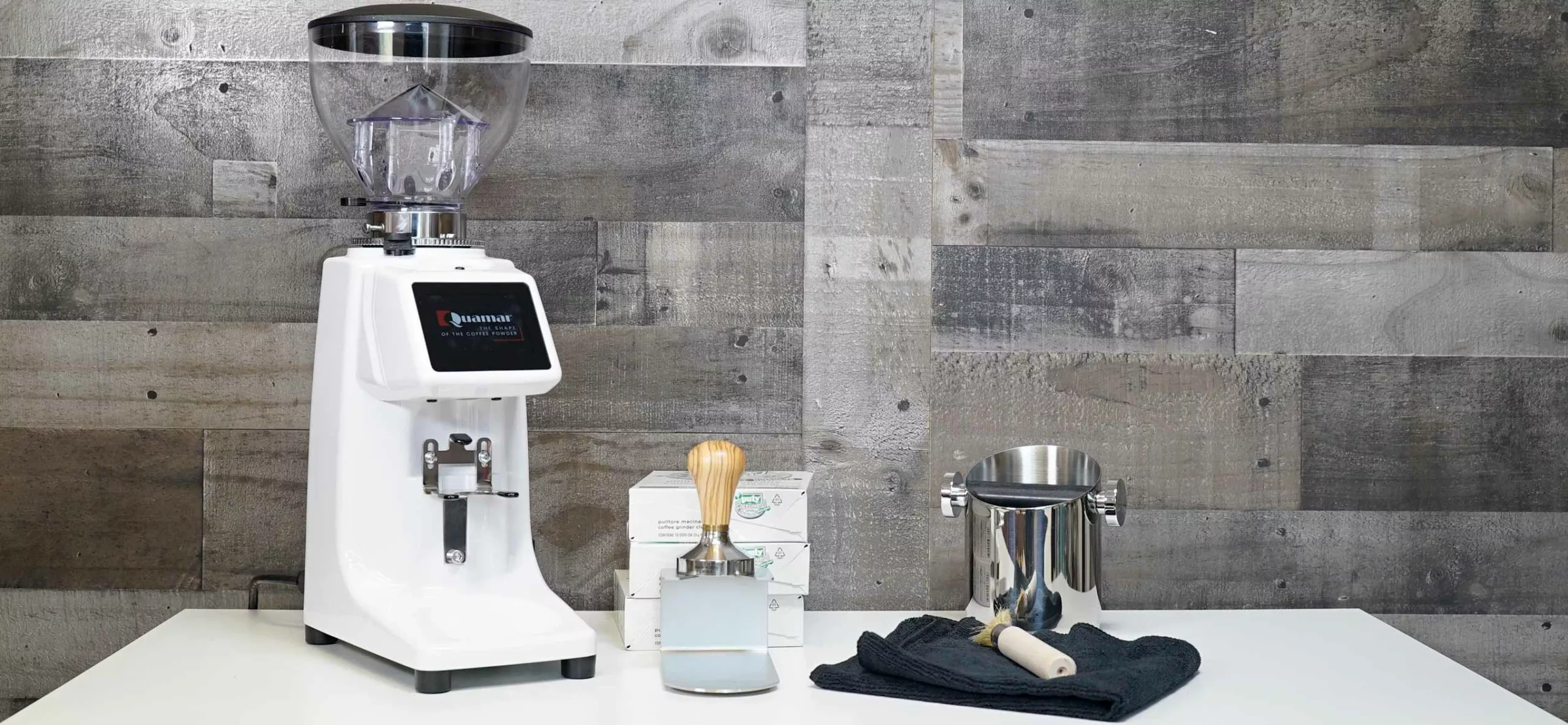 A Quamar M80TP Espresso Coffee Grinder in white sits on a table beside a tamper, metal knock box, black towel, and box, with a gray wooden plank wall background. © 2025 1st-line.com