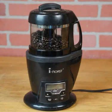 A SIDEWALK SALE i-Roast Home Coffee Roaster 110V (USED) (E110) in black sits on a wooden surface with dark roasted beans, set against a brick wall—ideal for coffee enthusiasts seeking their next great find. © 2025 1st-line.com