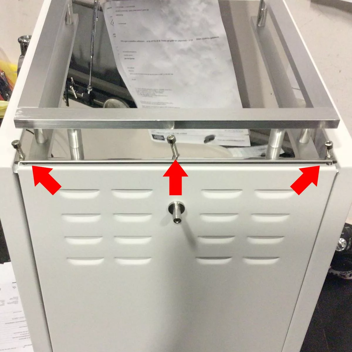A white metal cabinet with a partially open top lid and three large red arrows highlighting bolts on the front edge, key for changing the side panel. Papers are visible in the background. Compatible with Bellezza Bellona models. © 2025 1st-line.com