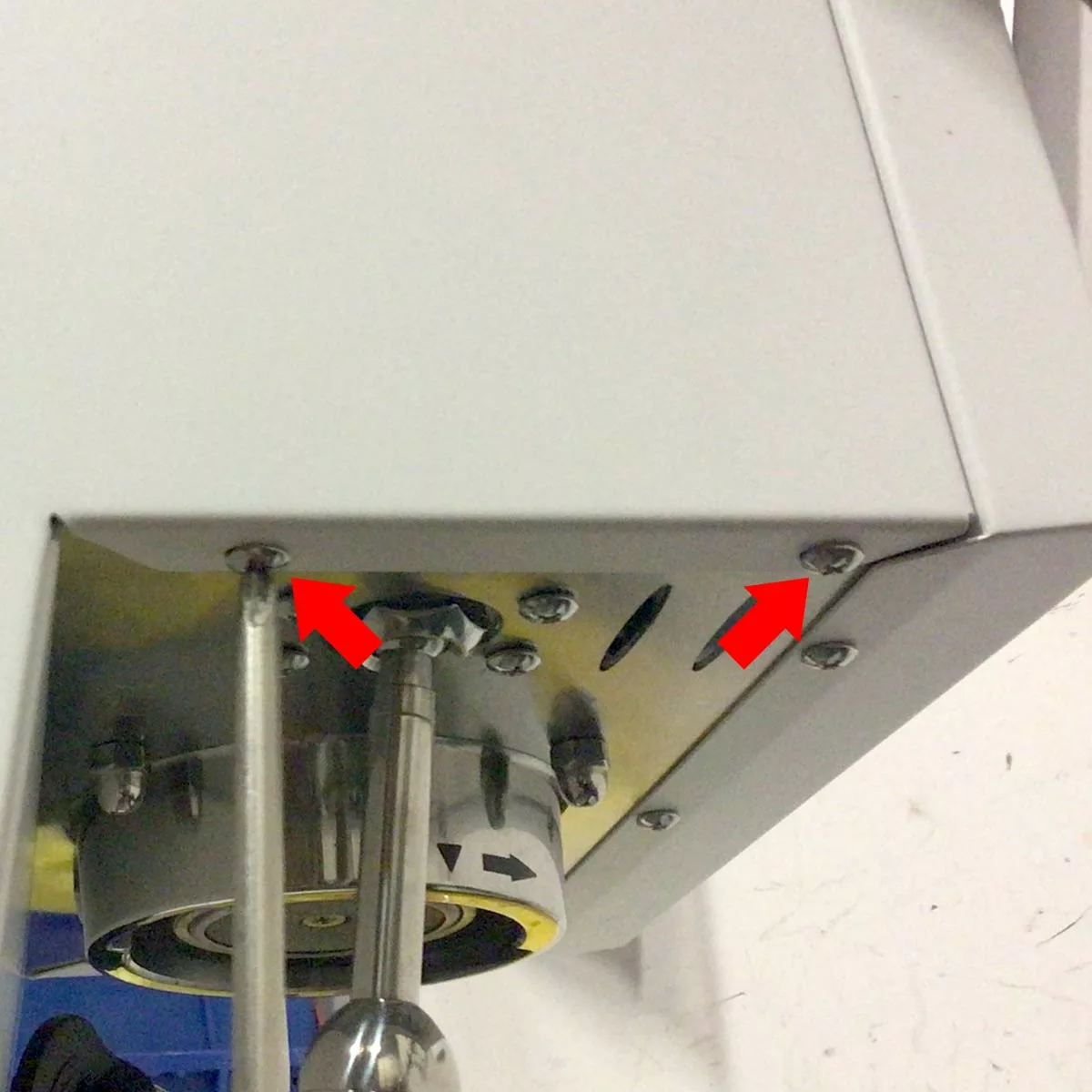 Close-up of a Bellezza Bellona machine corner with two red arrows pointing at screws, indicating where to unscrew for changing side panel. Various metallic components and a white surface are visible in the background. © 2025 1st-line.com