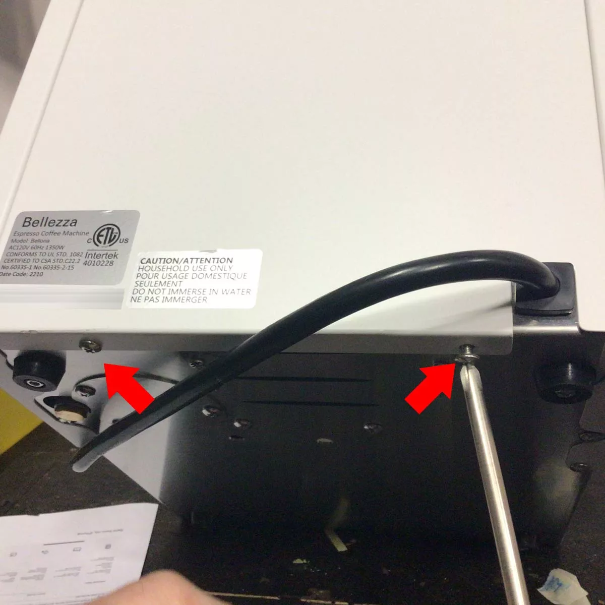 Close-up of the back of a Bellezza Bellona espresso machine: two red arrows highlight screws being removed for changing the side panel; a power cord runs along the back, and a hand uses a screwdriver to loosen one screw. © 2025 1st-line.com