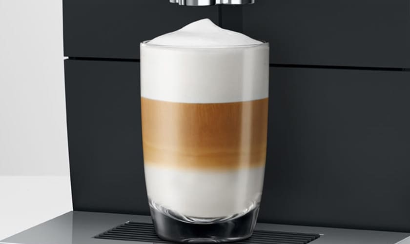 A clear glass of layered latte macchiato sits in front of the Jura ENA 8 Super-Automatic Espresso Machine, showcasing distinct layers of milk, espresso, and foam. © 2025 1st-line.com