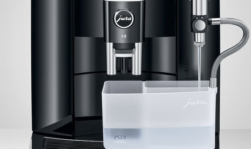 A close-up of the Jura E8 Super-Automatic Espresso Machine displays its transparent water container, partially filled, with a metal tube extending inside. © 2025 1st-line.com