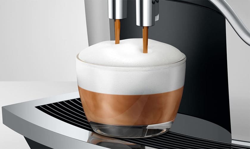 A glass cup is filled with cappuccino from the Jura E6 Super-Automatic Espresso Machine, featuring rich espresso pouring from two spouts and topped with creamy milk foam. © 2025 1st-line.com
