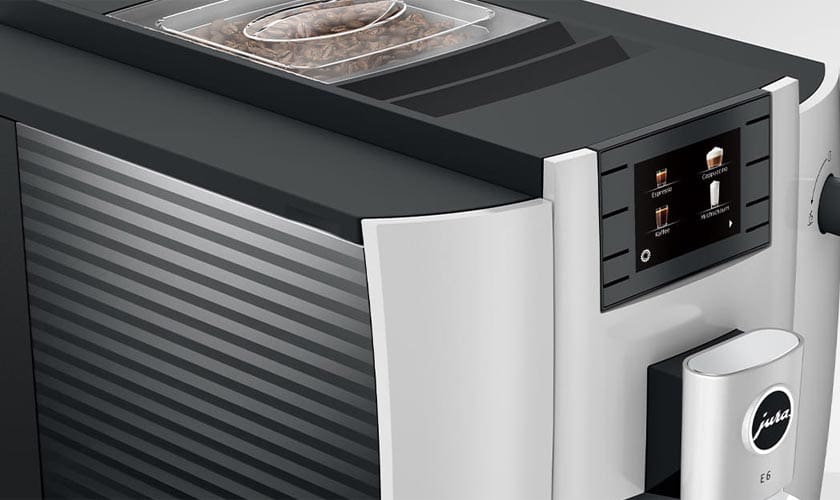 Close-up of the Jura E6 Super-Automatic Espresso Machine, showcasing its display screen for selecting drinks and a transparent top-mounted bean container filled with coffee beans. © 2025 1st-line.com