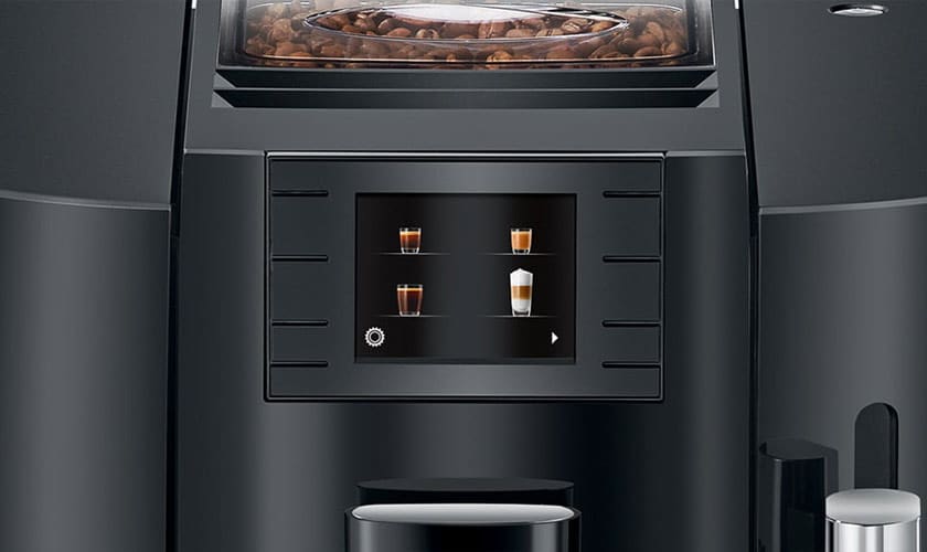 Close-up of the Jura E8 Super-Automatic Espresso Machines touch screen, displaying four coffee drink options. Coffee beans are visible in the compartment above. The machine features a sleek black modern design. © 2025 1st-line.com