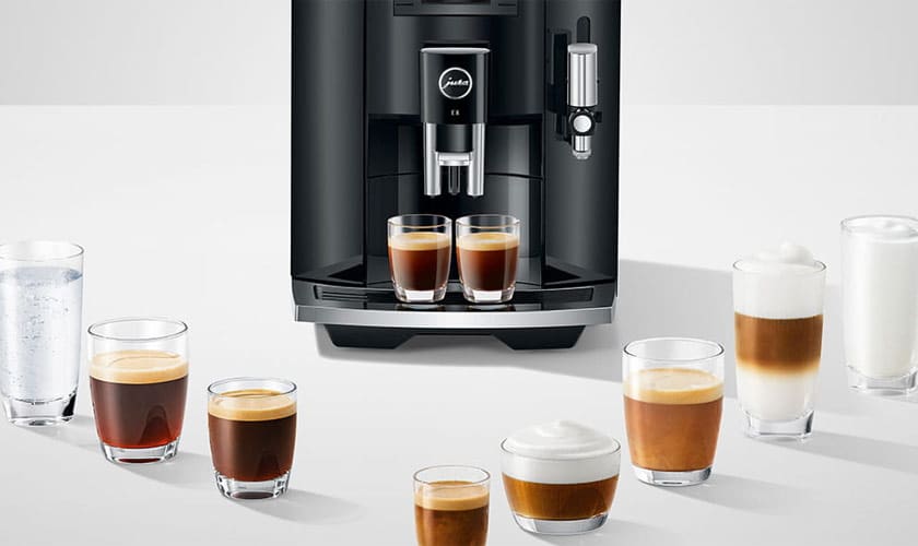 The black Jura E8 Super-Automatic Espresso Machine dispenses espresso into two small glasses, surrounded by lattes, cappuccinos, and a glass of water in clear cups on a white surface. © 2025 1st-line.com