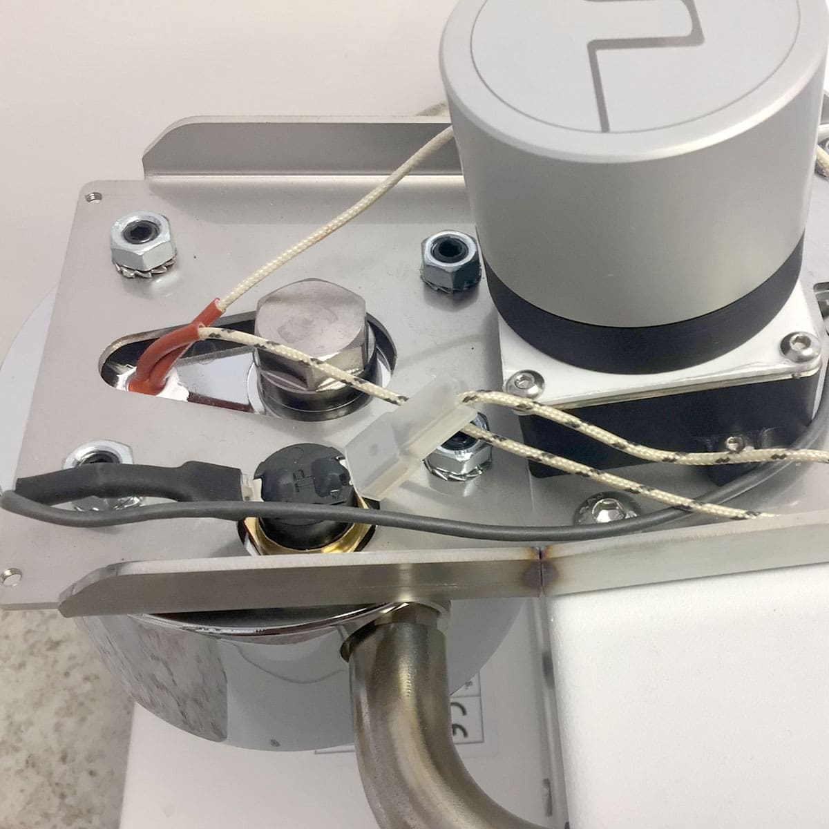 A close-up of the Nurri R-Type SB (HX) Espresso Machine shows wires, connectors, and cylindrical parts on a white and metallic base, set against a plain white wall and part of a floor. © 2025 1st-line.com