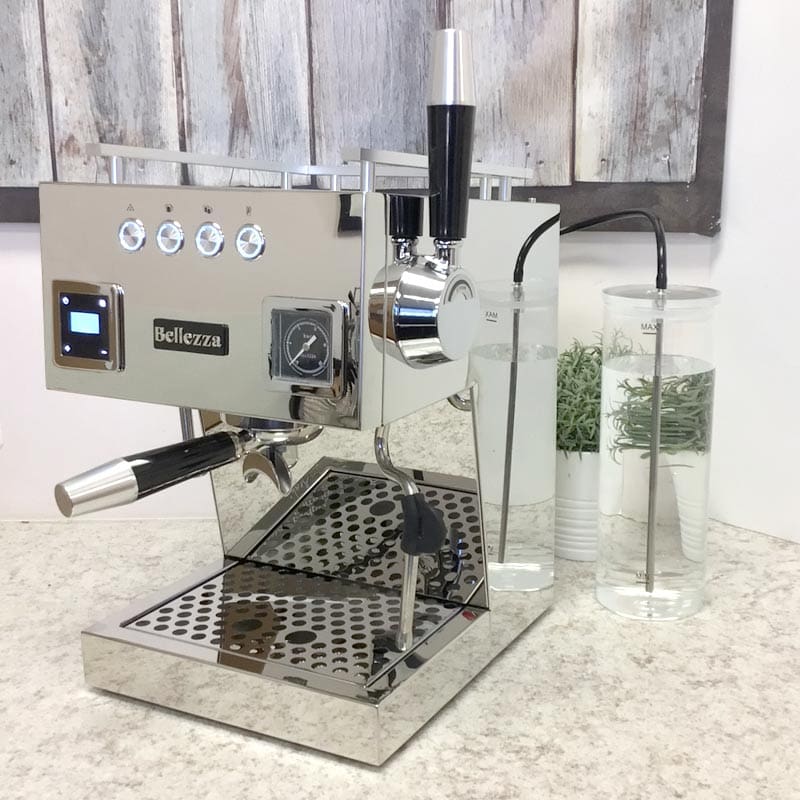 An OPEN BOX - Bellezza Bellona Dual Boiler Espresso Machine (C403) with a steam wand and gauges sits on a light countertop, beside two tall glass containers with water and herbs. A rustic wooden panel hangs on the wall behind. © 2025 1st-line.com