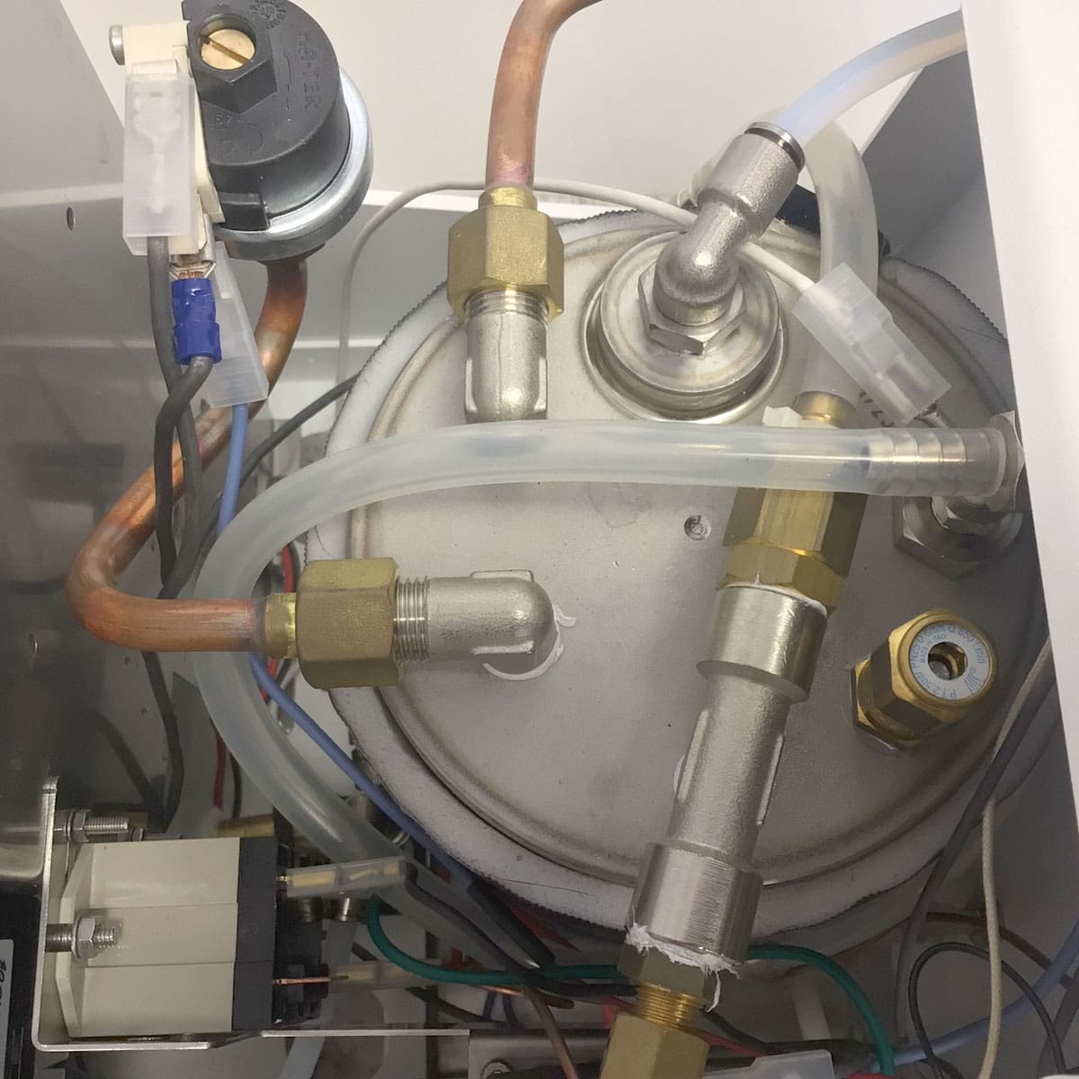 Close-up view of the internal components of a Nurri R-Type SB (HX) Espresso Machine, highlighting its metallic pipes, brass fittings, tubing, wiring, and a round metal tank—key parts of its advanced boiler system. © 2025 1st-line.com