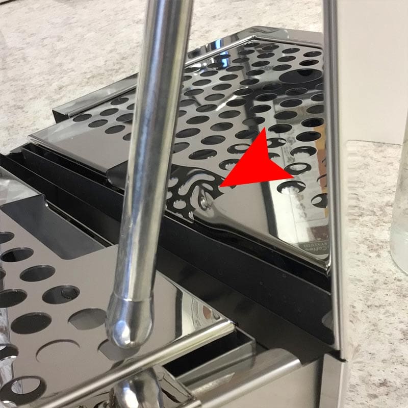 The OPEN BOX - Bellezza Bellona Dual Boiler Espresso Machine (C403) chrome drip tray displays circular holes, a metal arm in the foreground, and a red arrow marking a swirl-shaped scratch on the trays surface. © 2025 1st-line.com