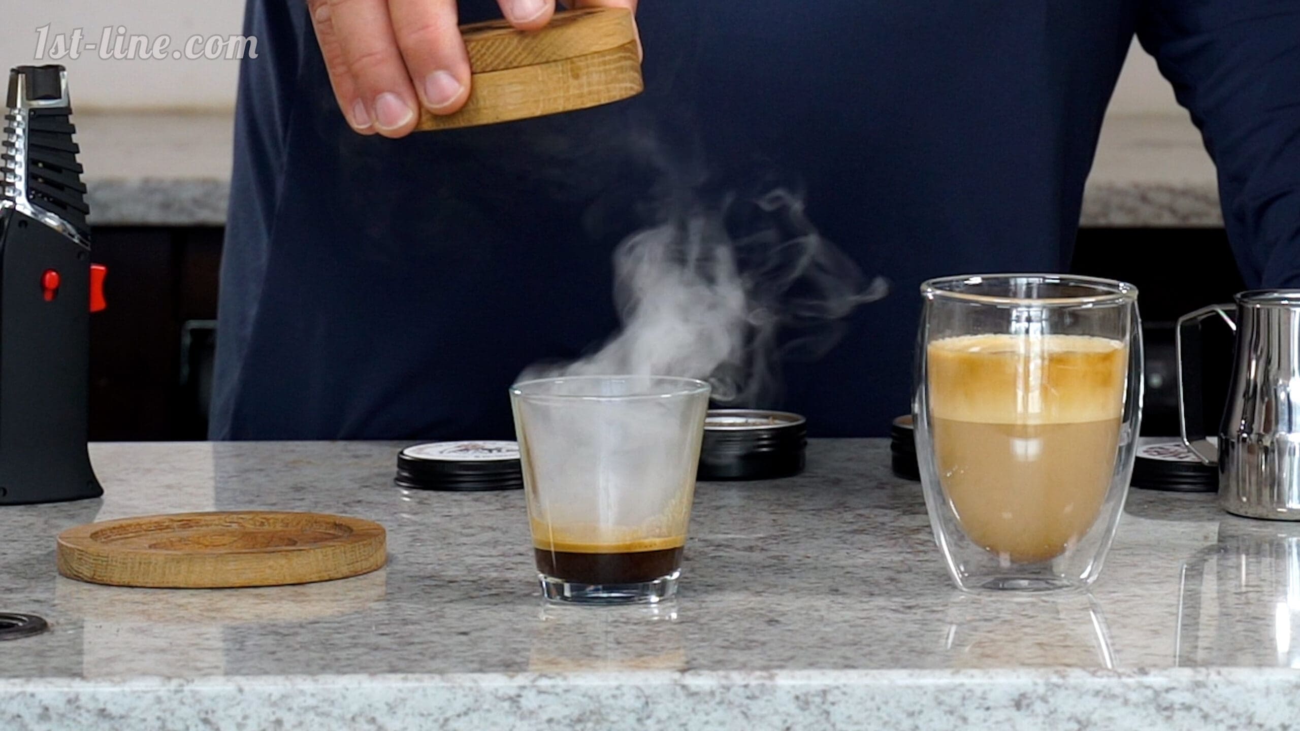 Using the Java Jim Espresso Smoker Kit, someone holds a wooden lid above a steaming glass of espresso on a countertop beside a coffee with milk and assorted accessories. © 2025 1st-line.com