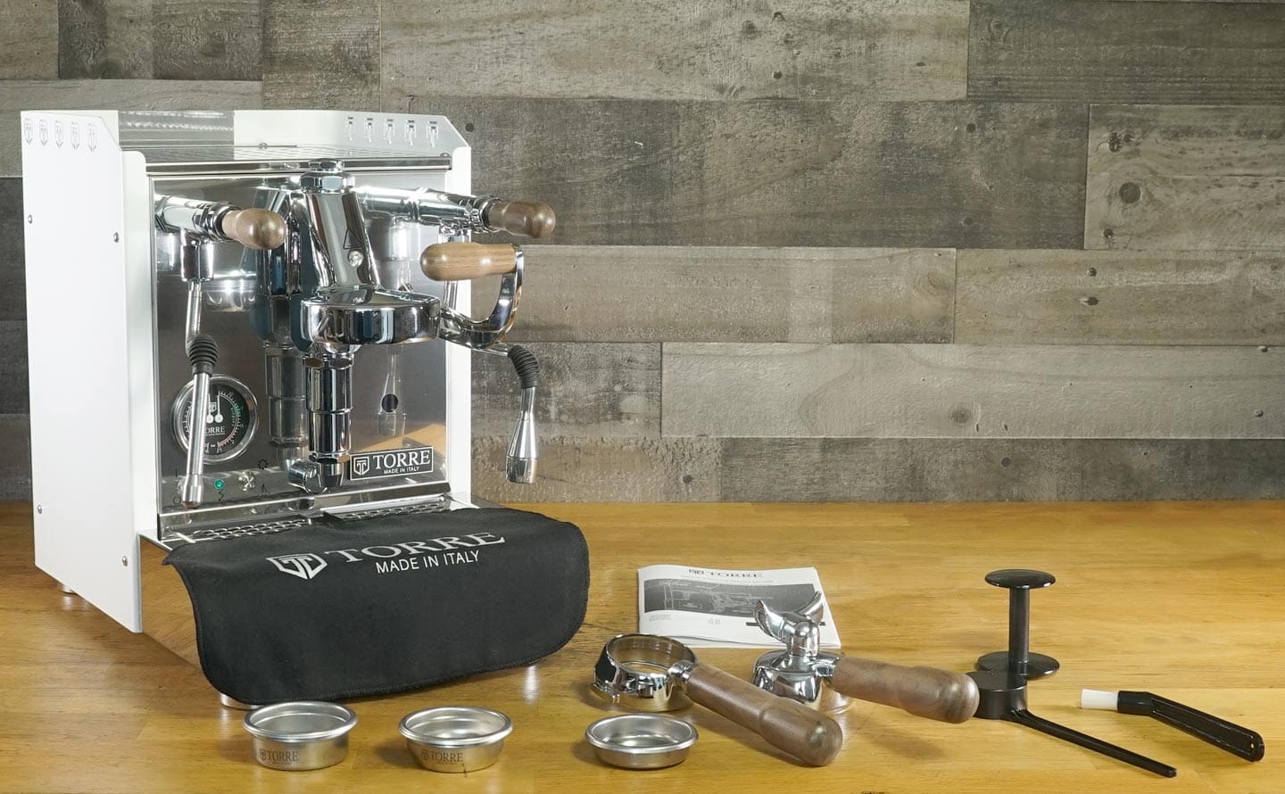 A Torre Alfredo TSP HX Espresso Machine with wooden handles rests on a wooden counter, accompanied by portafilters, filter baskets, a black tamper, brush, manual, and a branded Torre Alfredo cloth cover. © 2025 1st-line.com