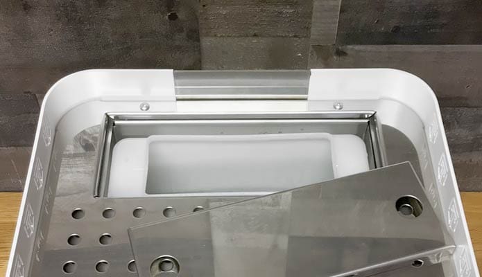 A white plastic container with a stainless steel insert and perforated tray, partially covered by a clear lid, sits on a wooden surface next to the Torre Alfredo TSP HX Espresso Machine against a grey tiled wall. © 2025 1st-line.com