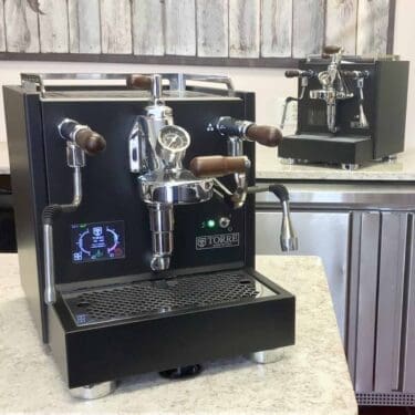 An open box SIDEWALK SALE Torre Peppina EVO Premium Dual Boiler Espresso Machine sits on a light countertop, featuring wooden handles and a pressure gauge, with another similar machine visible in the background. © 2025 1st-line.com