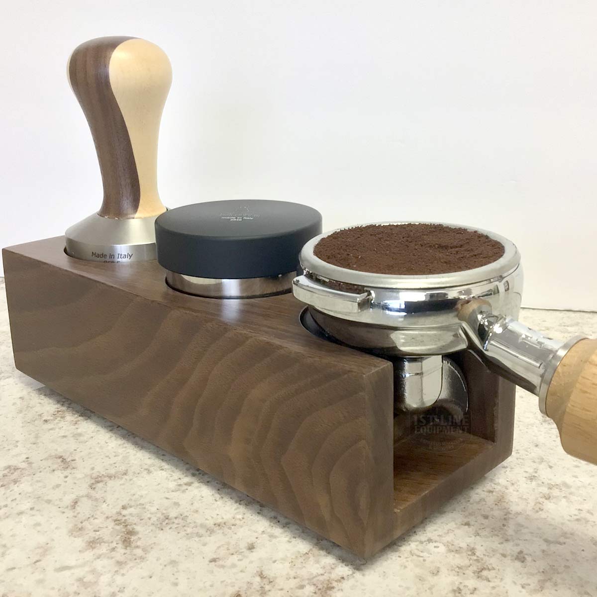 The Eureka Walnut Tamping Station - double housing holds a coffee tamper, distributor, and portafilter filled with ground coffee on a light countertop. © 2025 1st-line.com