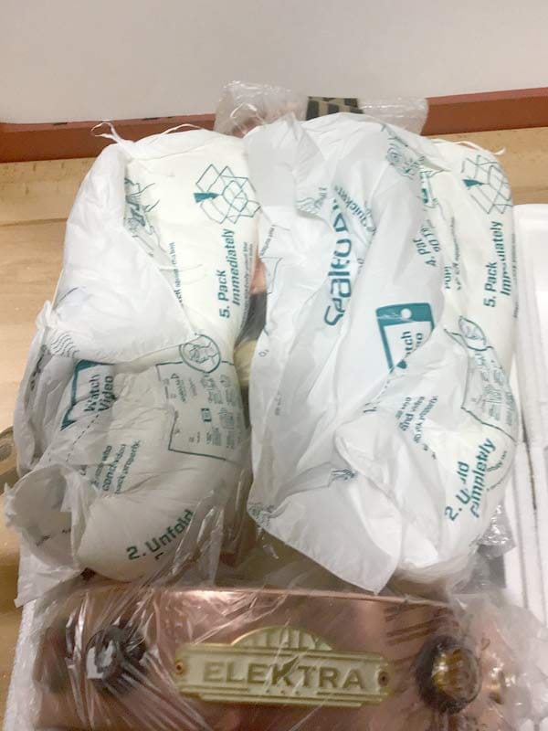 A partially unpacked Elektra Semi-Automatica espresso machine, covered in protective plastic and surrounded by white air cushion packaging, awaits repackaging. The brand name Elektra is visible on a metal plate. © 2025 1st-line.com