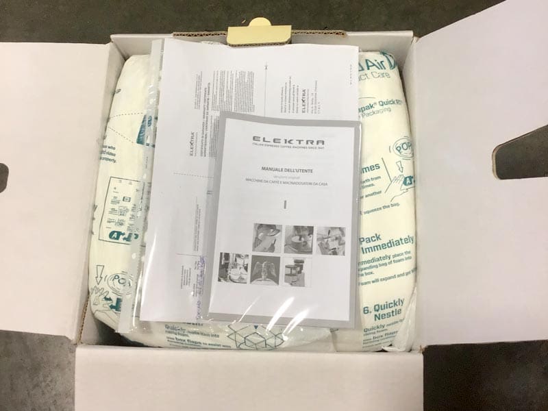 An opened white cardboard box contains sealed air packaging and a plastic-wrapped instruction manual labeled Elektra Semi-Automatica, with assembly instructions and diagrams visible on top, ready for repackaging. © 2025 1st-line.com