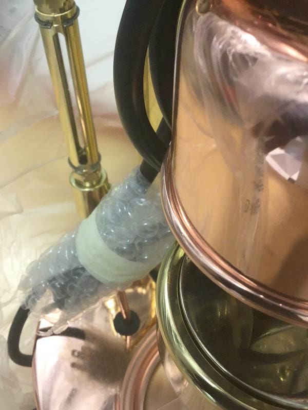 Close-up of a metallic object, possibly an Elektra Semi-Automatica, with brass and copper parts. One section is wrapped in bubble wrap and plastic for repackaging. Black electrical cords are also visible. © 2025 1st-line.com