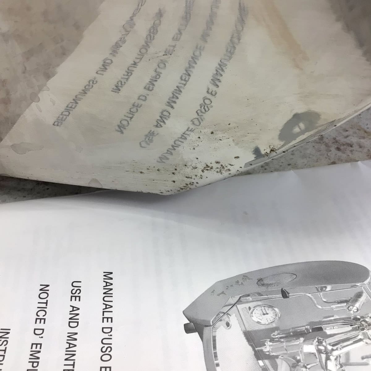 A close-up of a SIDEWALK SALE - Isomac Mondiale HX Espresso Machine (E013) manual (Italian/English) sits beside a thin, semi-transparent sheet stained with dirt or grease, which partly covers the technical diagram on the manual. © 2025 1st-line.com