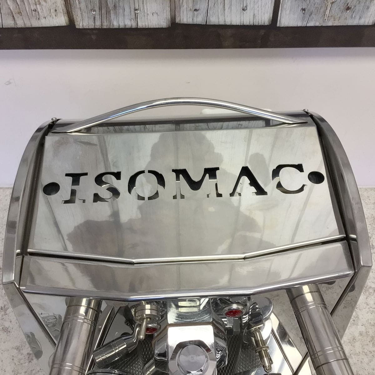 A close-up of the shiny SIDEWALK SALE - Isomac Mondiale HX Espresso Machine (E013) with ISOMAC cut-out on its reflective top, visible pipes and buttons below—set against a wooden wall for an eye-catching display. © 2025 1st-line.com