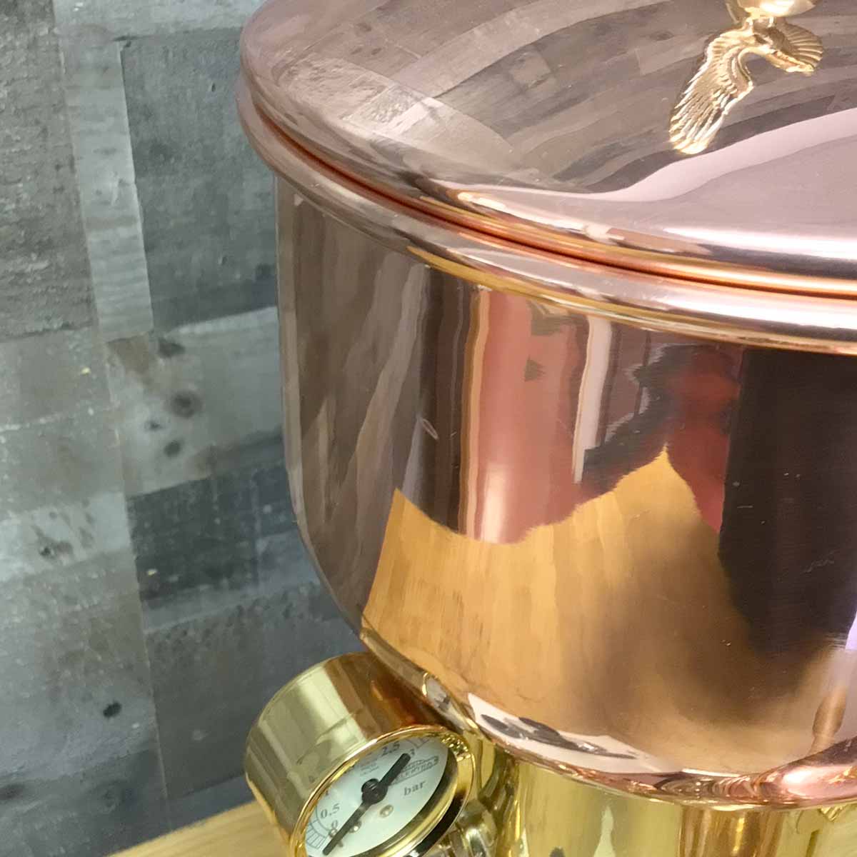 A close-up of the SIDEWALK SALE - Elektra SX Microcasa Semiautomatica Espresso Machine (RETURNED & REFURBISHED, E211) shows its shiny copper design and round lid with pressure gauge, set against a gray wooden background. © 2025 1st-line.com