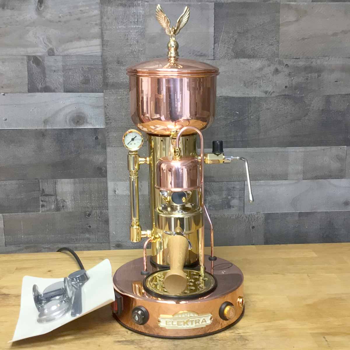 A SIDEWALK SALE - Elektra SX Microcasa Semiautomatica Espresso Machine RETURNED & REFURBISHED (E211) with an eagle finial, wooden handle, and pressure gauge sits on a wooden surface, its power cord neatly coiled on a white tray nearby. © 2025 1st-line.com