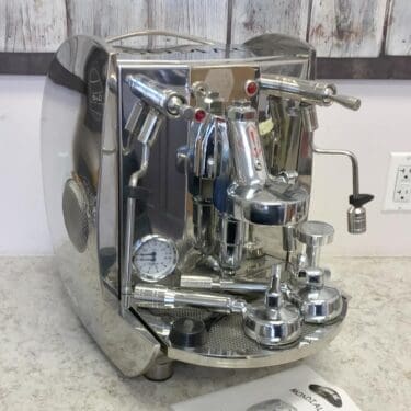 A SIDEWALK SALE - Isomac Mondiale HX Espresso Machine (E013) with stainless steel finish, levers, dials, and steam wand sits on a countertop; its user manual is in front and a rustic wooden wall is in the background. © 2025 1st-line.com