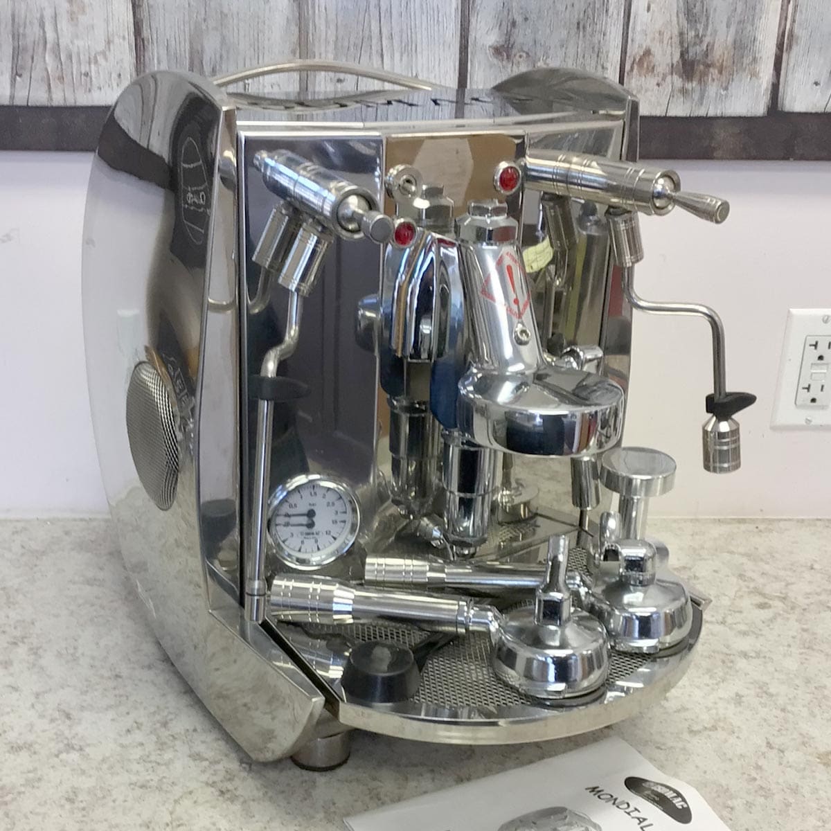 A SIDEWALK SALE - Isomac Mondiale HX Espresso Machine (E013) with stainless steel finish, levers, dials, and steam wand sits on a countertop; its user manual is in front and a rustic wooden wall is in the background. © 2025 1st-line.com