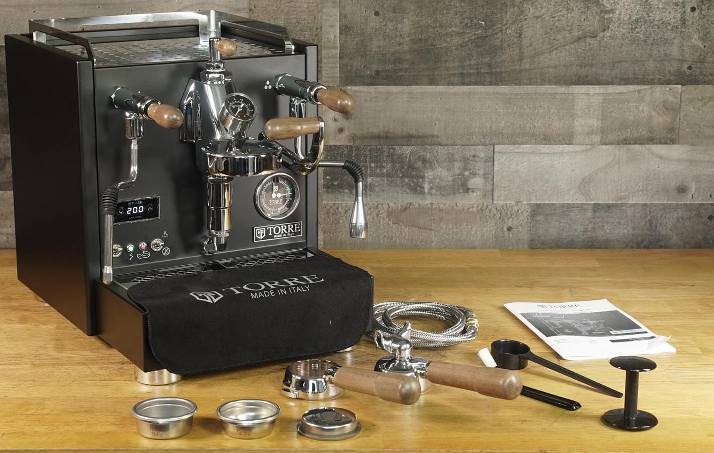 A Torre Mariuccia Dual Boiler Espresso Machine w/ Flow Control with wood accents sits on a wooden counter, surrounded by portafilters, baskets, tamper, metal hose, black cloth, and an instruction manual for precise brewing. © 2025 1st-line.com