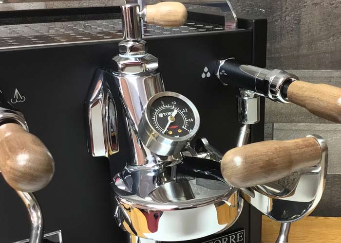 Close-up of the Torre Mariuccia Dual Boiler Espresso Machine w/ Flow Control, featuring shiny metal, wooden handles, and a central pressure gauge showcasing its flow control function against a dark background. © 2025 1st-line.com