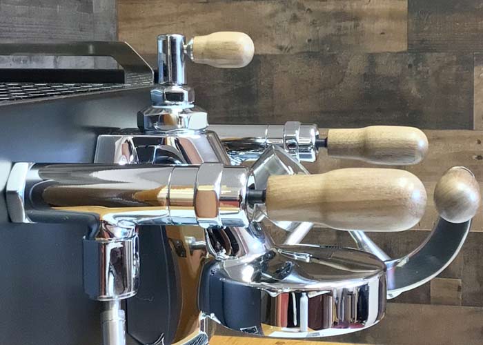 Close-up of the Torre Mariuccia Dual Boiler Espresso Machine w/ Flow Control, featuring shiny metal, wooden handles, portafilters, steam wand, and advanced flow control, set against a wood-patterned background. © 2025 1st-line.com