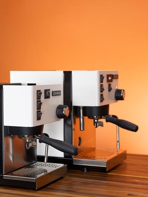 Two modern white espresso machines, including the Rancilio Silvia Pro X and Silvia Classic, with black handles are placed side by side on a wooden counter, set against an orange background. © 2025 1st-line.com