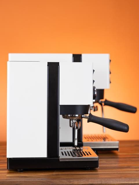 Two modern, white espresso machines, including the Rancilio Silvia Pro X, with black accents are placed side by side on a wooden surface, against an orange background. © 2025 1st-line.com