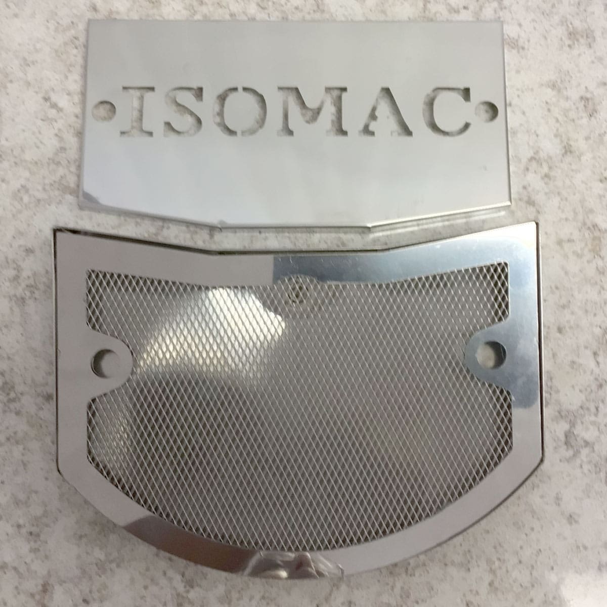 Two shiny metal parts are displayed on a stone surface for the SIDEWALK SALE - Isomac Mondiale HX Espresso Machine (E013): one with ISOMAC cut out, the other a mesh with a solid frame and two screw holes. © 2025 1st-line.com