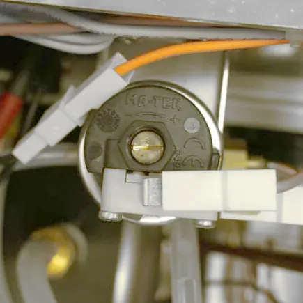 Close-up view of an electrical component labeled HA-TER on its circular, black plastic surface—part of the Elektra Verve steam boiler system. Orange and white wires are attached, with metal and plastic parts visible around it for precise pressure adjustment. © 2025 1st-line.com