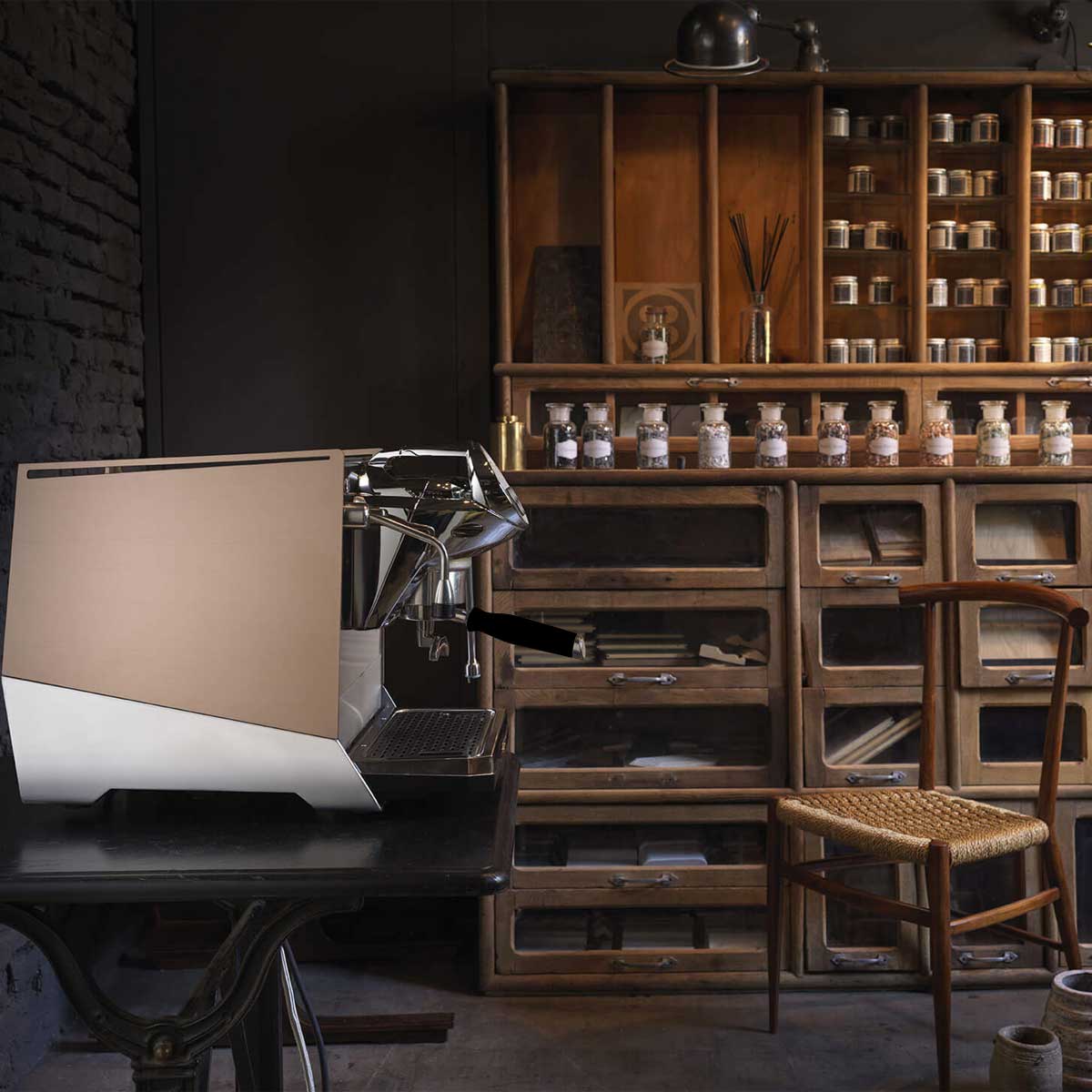 A Faema Faemina Dual Boiler Espresso Machine with AUTOSTEAM sits on a dark wooden table in a rustic room, surrounded by shelves of jars, wooden drawers, and a woven chair beside the table. © 2025 1st-line.com