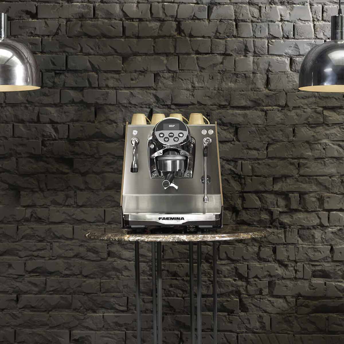 A Faema Faemina Dual Boiler Espresso Machine with AUTOSTEAM sits on a small round marble table against a dark gray brick wall, illuminated by two industrial-style pendant lights above. © 2025 1st-line.com