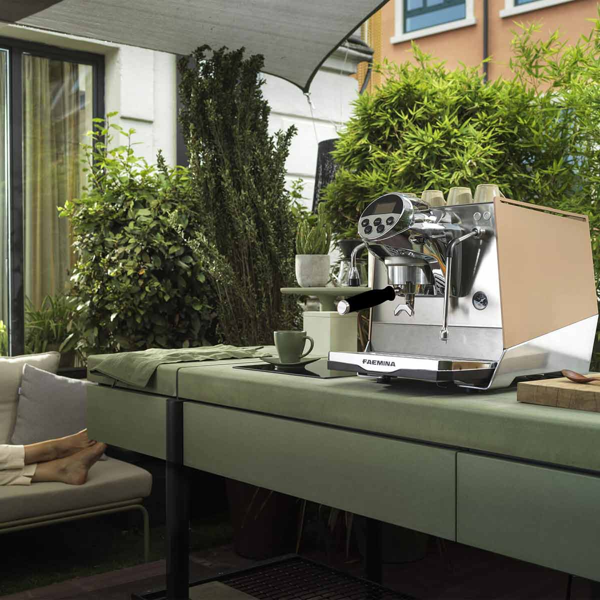 A Faema Faemina Dual Boiler Espresso Machine with AUTOSTEAM sits on an outdoor counter beside a green cup and saucer, surrounded by lush plants, while someone relaxes on a sofa with their feet visible in the background. © 2025 1st-line.com