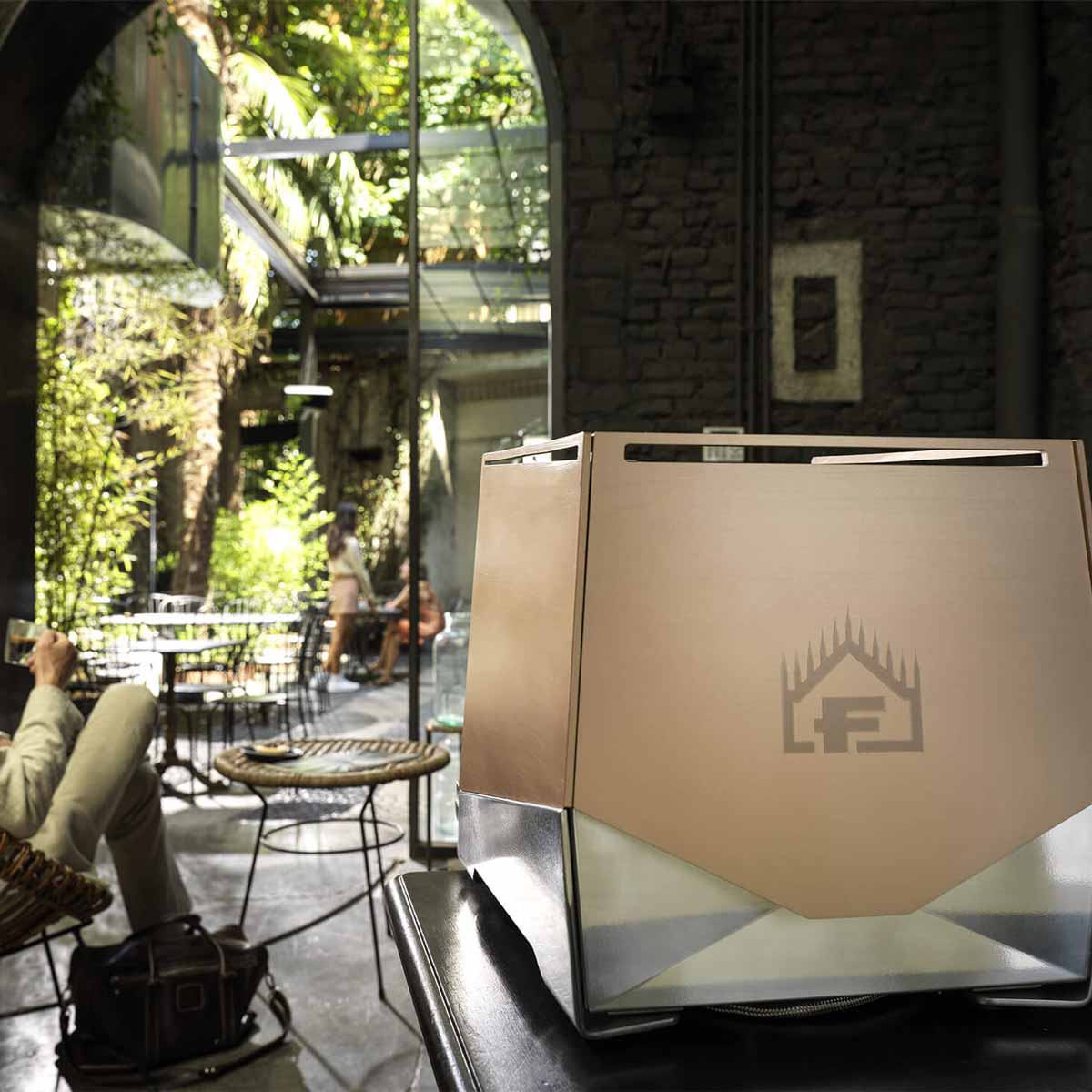 A gold Faema Faemina Dual Boiler Espresso Machine with AUTOSTEAM and geometric design sits on a table in a stylish café with large windows and stone walls. Someone relaxes nearby, enjoying this elegant espresso machine. © 2025 1st-line.com