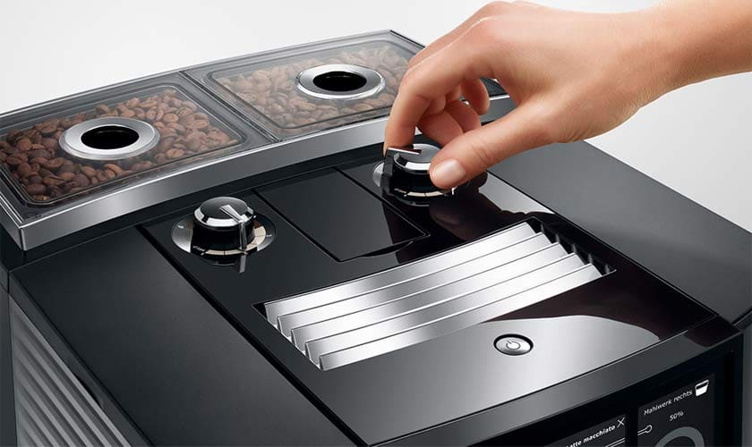A hand adjusts a dial on the Jura J8 Twin Super-Automatic Espresso Machine, which features two transparent bean containers on top filled with coffee beans. © 2025 1st-line.com