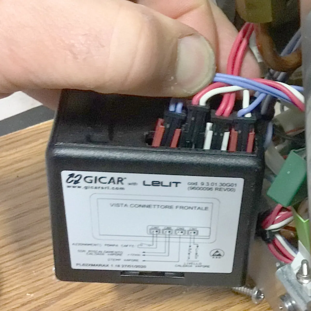 A hand connects multicolored wires to a black Gicar Brain Unit control box labeled &ldquo;LELIT&rdquo; with a wiring diagram on the front panel. The box, part of the Lelit Mara X espresso machine, is installed in an electronic device.