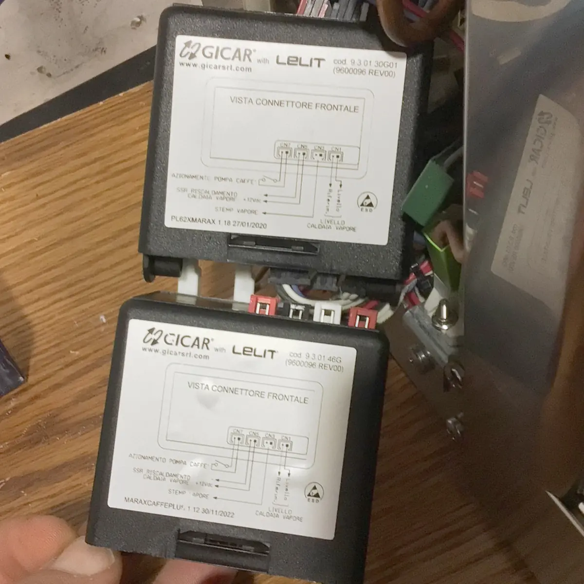 A hand holds a black electronic module labeled GICAR with LELIT near an identical Brain Unit installed in a Lelit Mara X machine, both displaying wiring diagrams and technical details. Wires are connected to the installed unit.