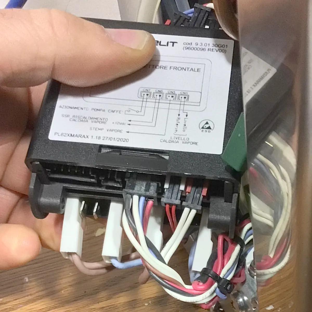 A hand holds a Brain Unit electronic control with various colored wires plugged into it. A diagram and technical information label are visible on the top, while a reflective metallic surface hints at its use in machines such as the Lelit Mara X.