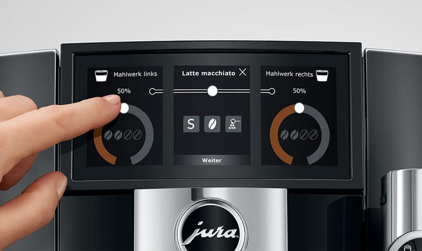 A hand touches the display of a Jura J8 Twin Super-Automatic Espresso Machine, adjusting coffee strength for a latte macchiato. The screen shows strength controls and coffee icons. © 2025 1st-line.com